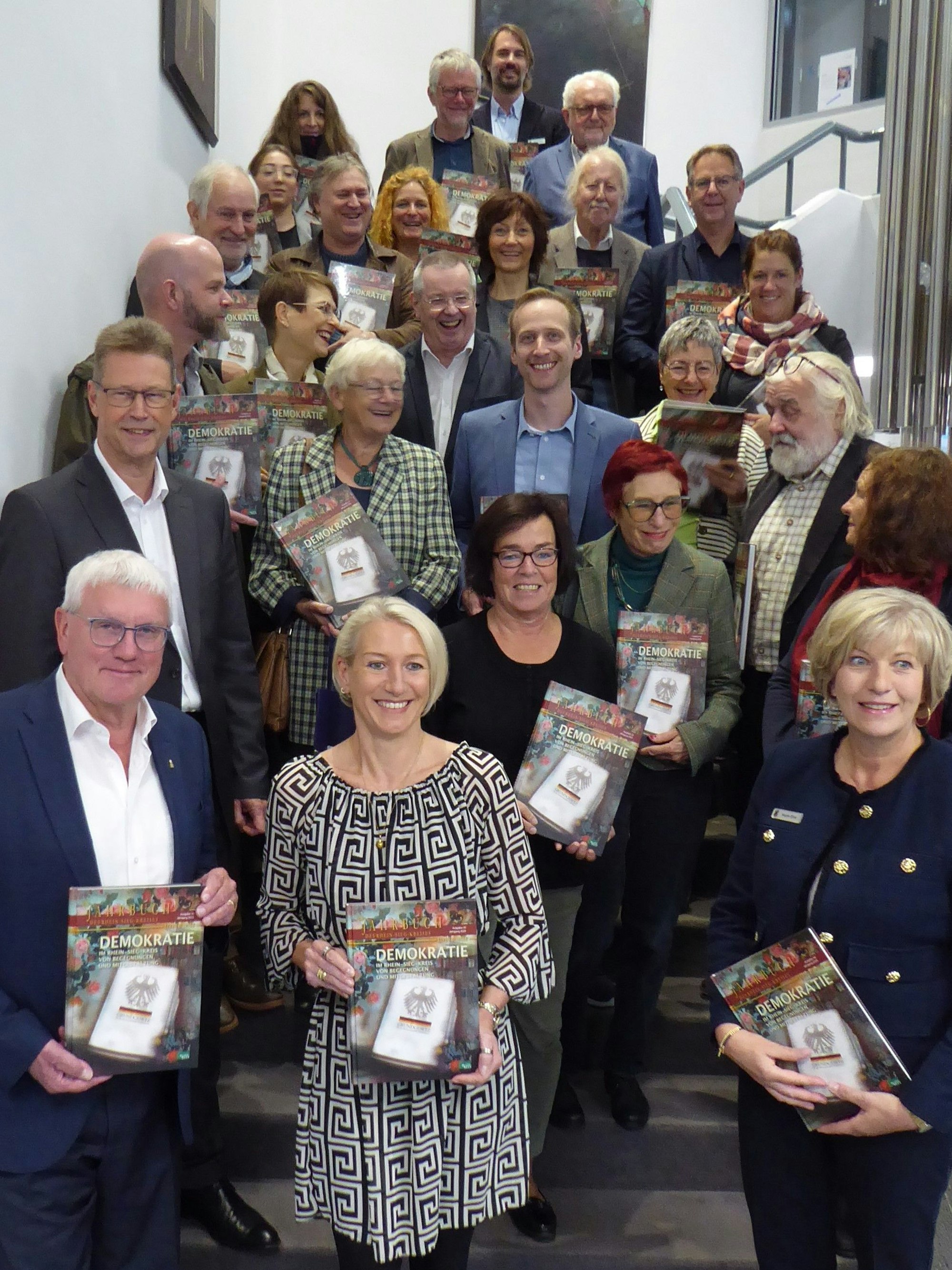 25 Männer und Frauen stehen auf einer Treppe und halten Bücher in den Händen.
