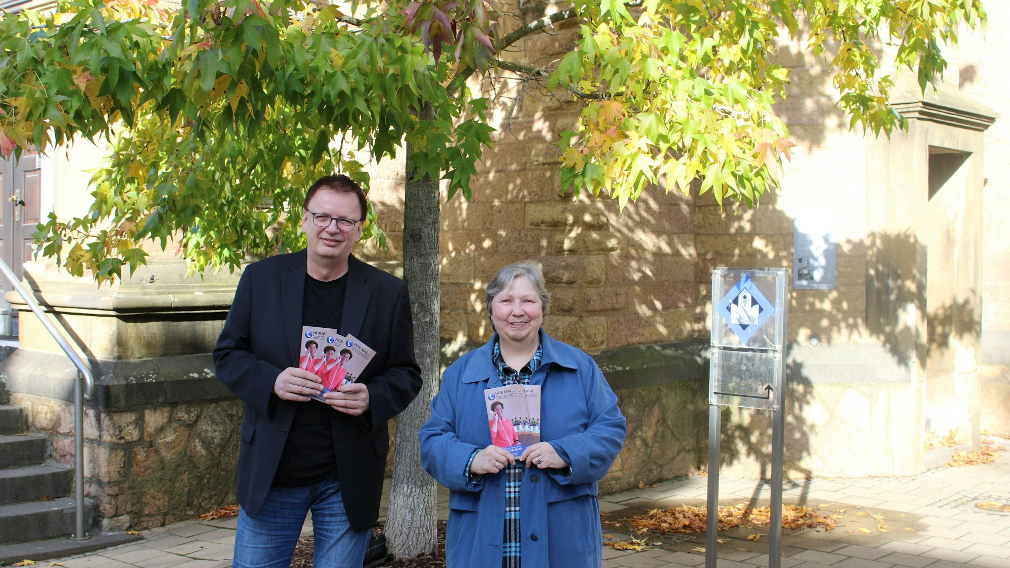 Ein Mann und eine Frau, Diakon Jens Schramm und Trudi Fleischhauer, stehen in Euskirchen vor einem Baum an einer Kirche. Sie halten Flyer in den Händen.