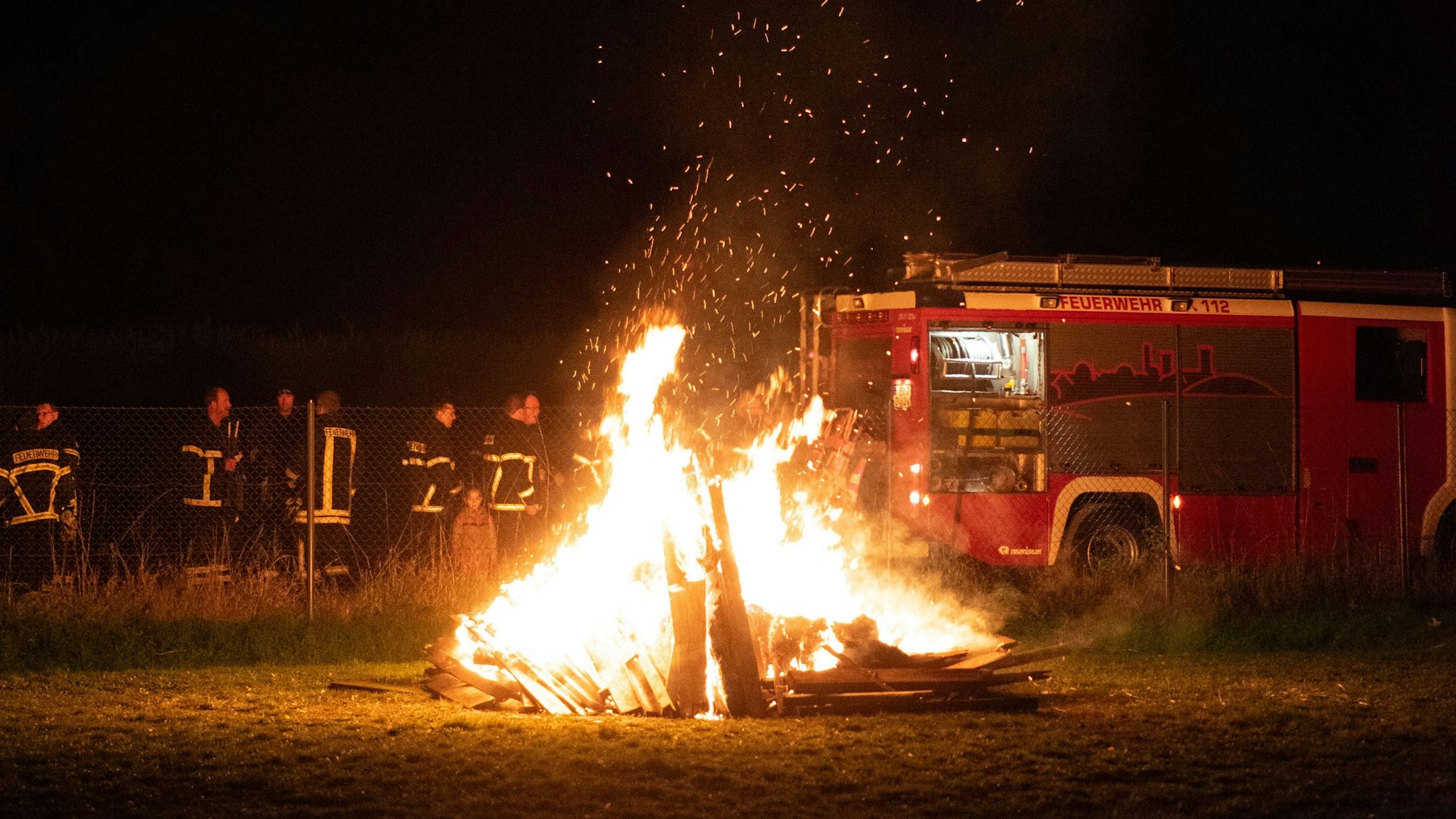 Ein St.-Martins-Feuer brennt auf einer Wiese. Dahinter sind Feuerwehrleute zu sehen.