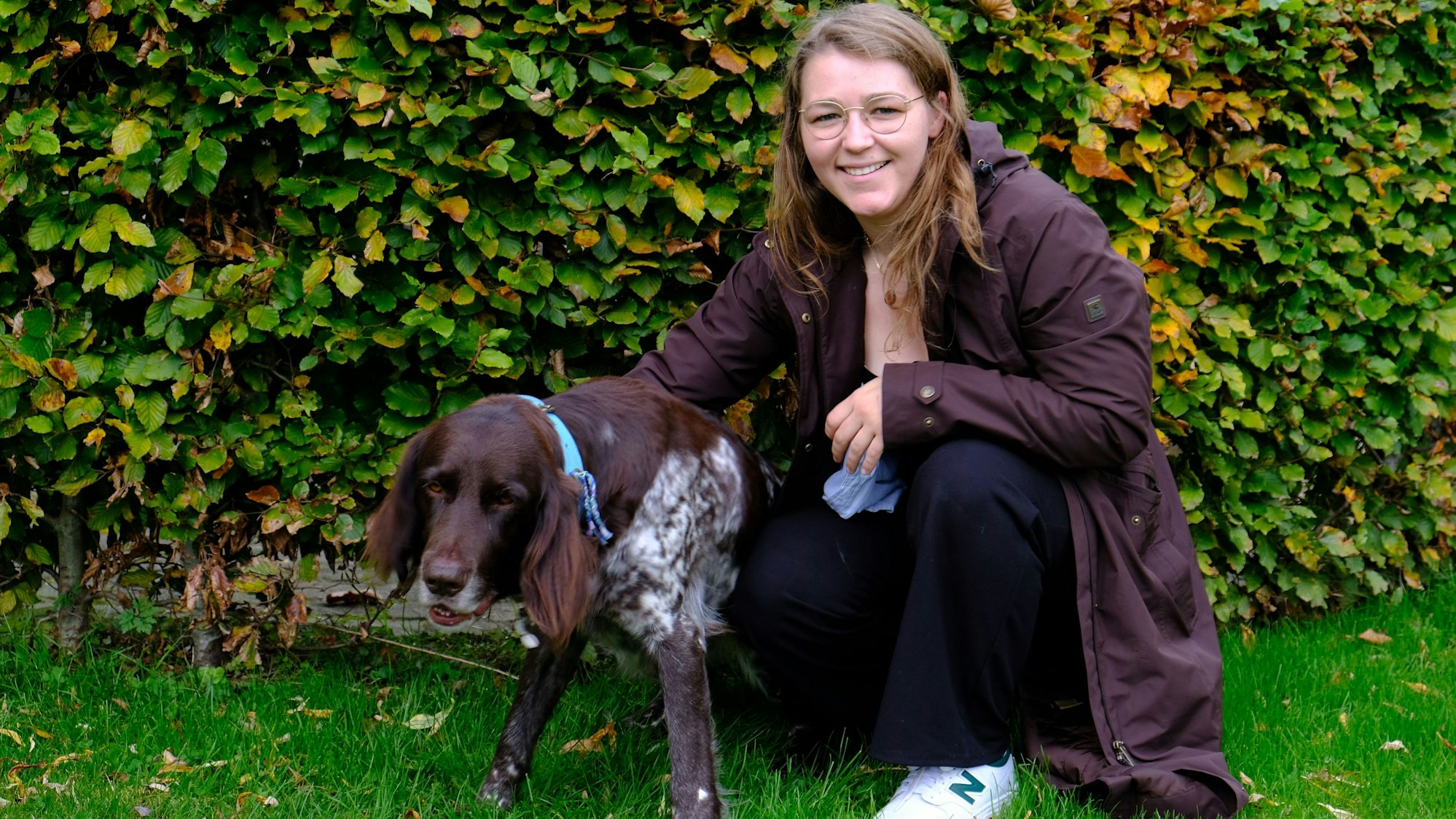 Tessa Riedling kniet mit Deutsch-Langhaar-Hündin Milly vor einer Hecke.