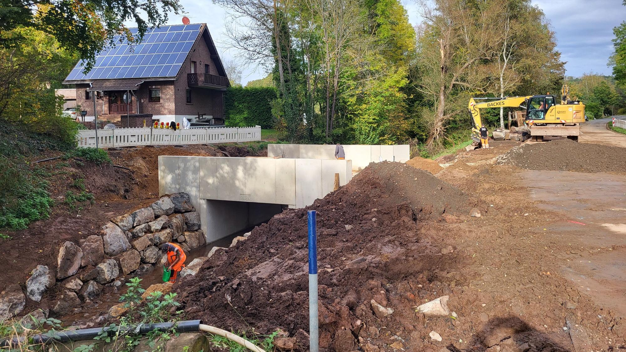 Das Bild zeigt eine neu gebaute Brücke, links ist sie mit Warnbarken abgesperrt, rechts sind Erdhügel und ein Bagger zu sehen.
