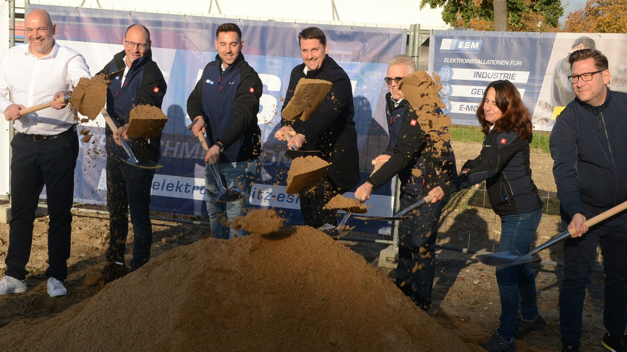 Sechs Männer und eine Frau werfen beim ersten Spatenstich auf einer Baustelle Erde in die Luft.