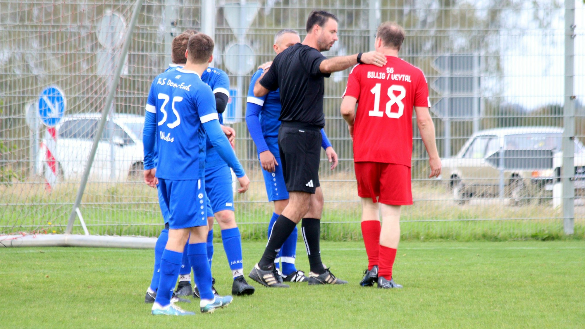 Ein Schiedsrichter im Gespräch mit zwei Spielern, denen er jeweils einen Arm auf die Schultern gelegt hat.