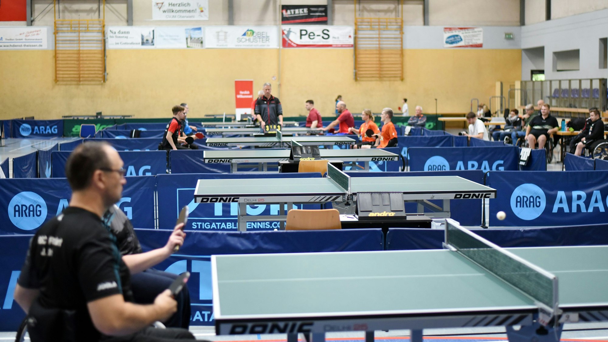 Blick in einen Sporthalle, in der Tischtennisplatten aufgebaut sind, an denen gespielt wird.