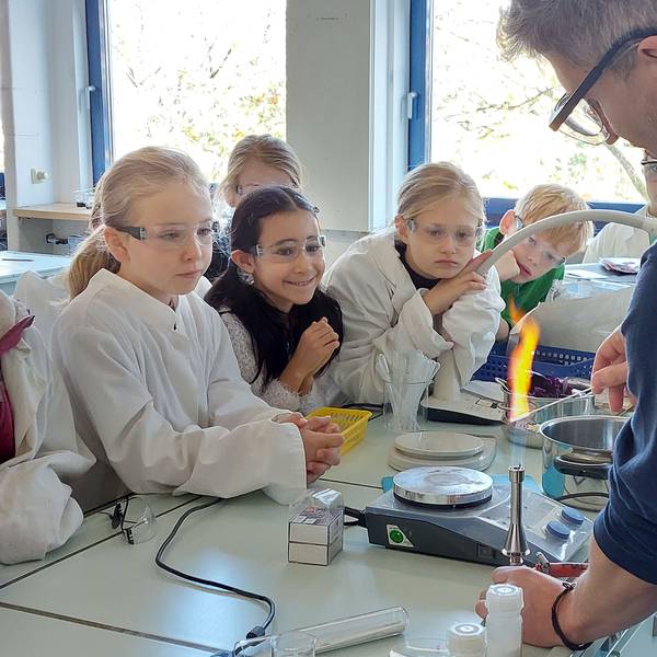 Jungen und Mädchen beobachten ein Experiment mit einem Bunsenbrenner. Die Kinder tragen Kittel und Schutzbrillen.