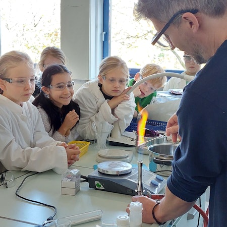 Jungen und Mädchen beobachten ein Experiment mit einem Bunsenbrenner. Die Kinder tragen Kittel und Schutzbrillen.