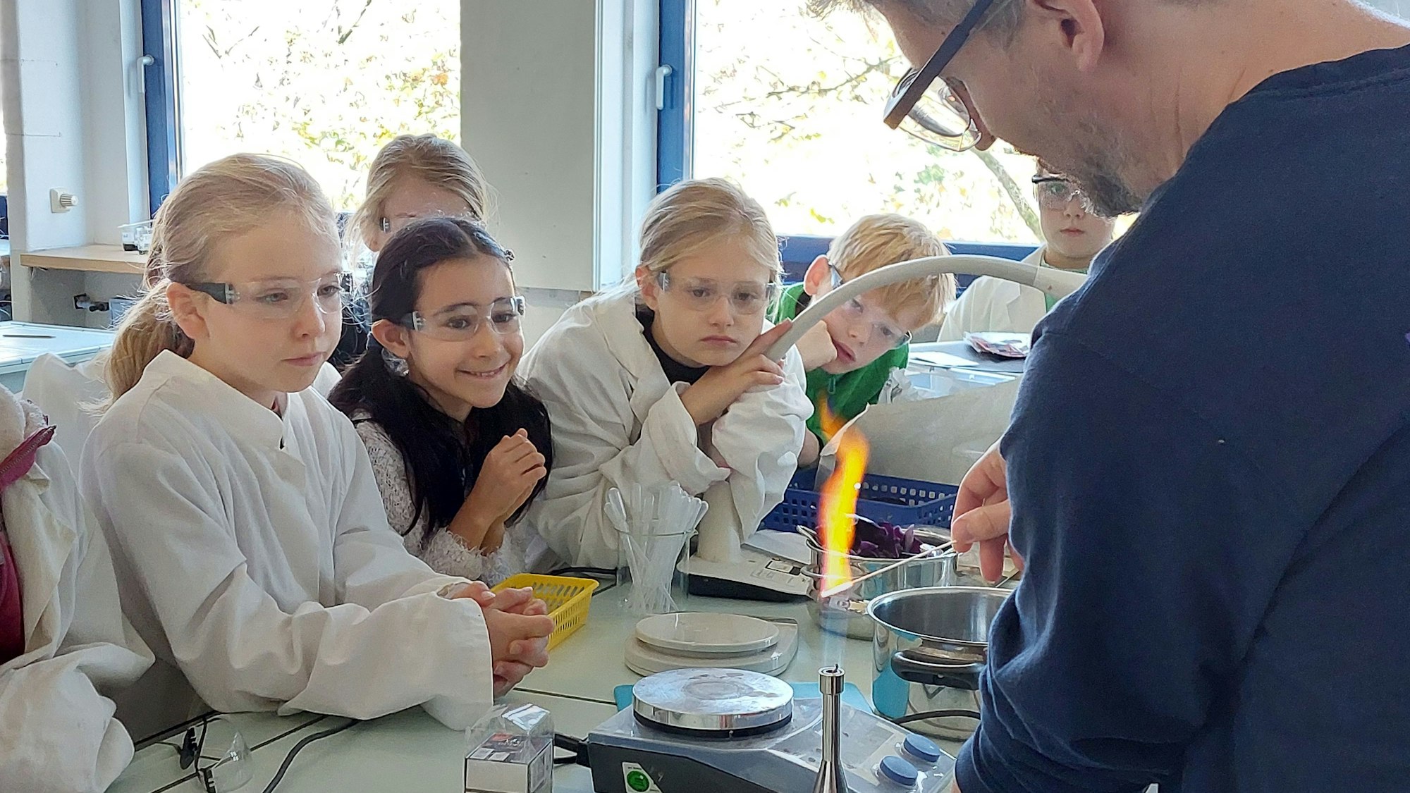 Jungen und Mädchen beobachten ein Experiment mit einem Bunsenbrenner. Die Kinder tragen Kittel und Schutzbrillen.