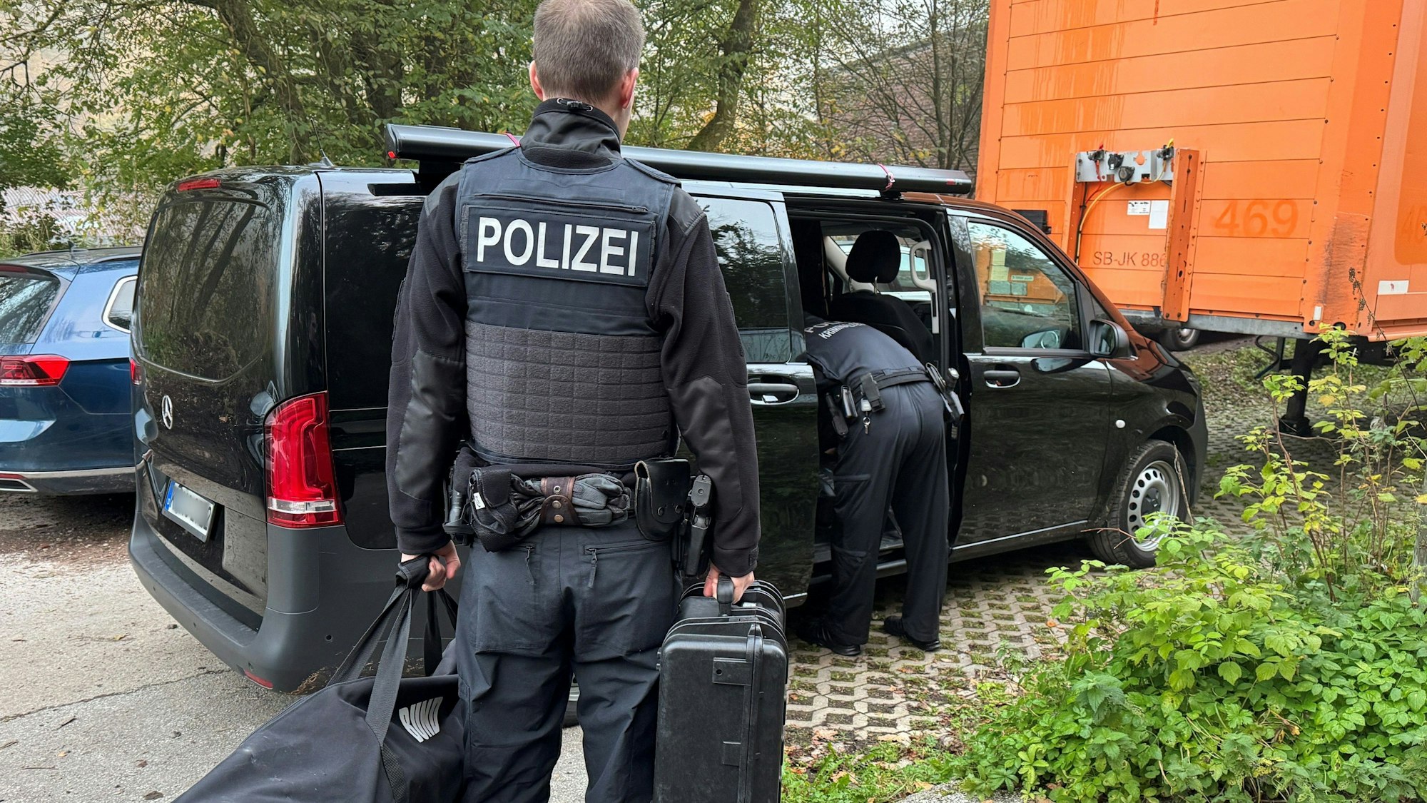 Ein Polizist steht mit einer Tasche und einem Koffer neben einem Wagen.