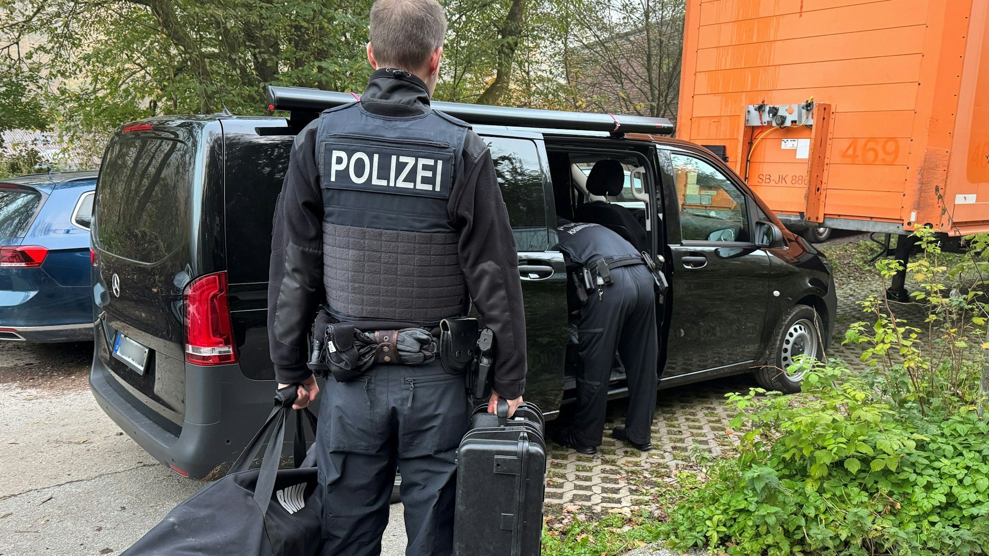 Ein Polizist steht mit einer Tasche und einem Koffer neben einem Wagen.