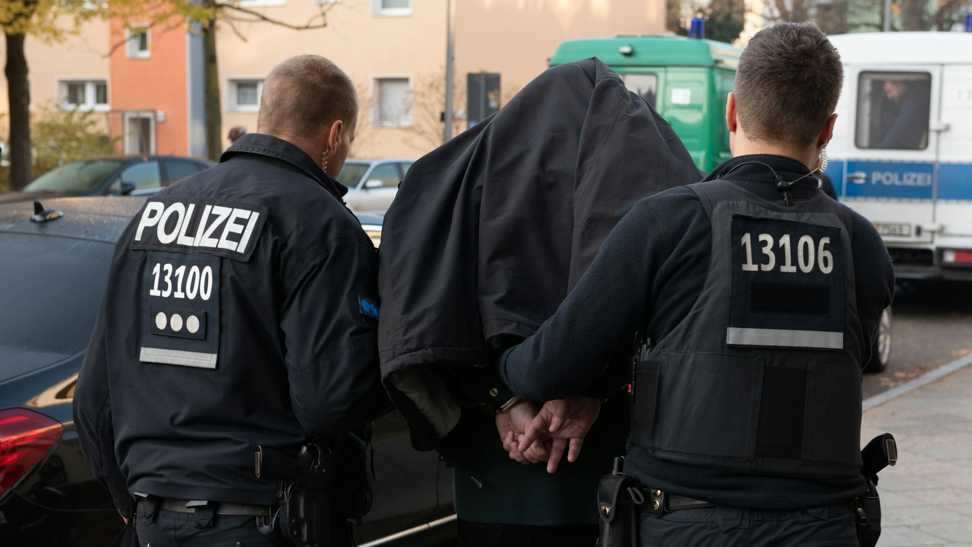 Zwei Polizisten führen eine Person in Handschellen ab, die eine Jacke über den Kopf trägt.