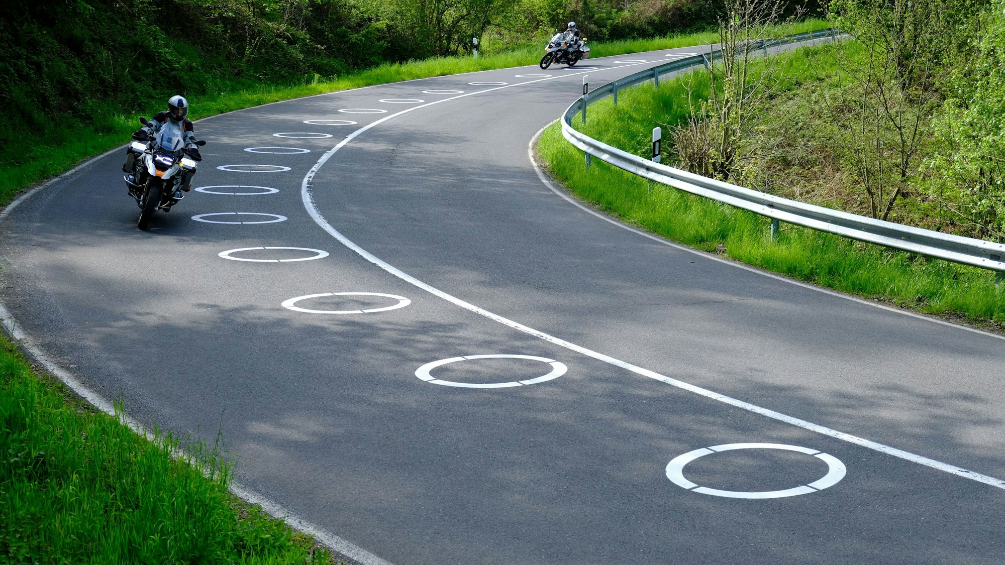 Auf einer Straße in der Eifel sind neben der Mittellinie ellipsenförmige Markierungen zu sehen. Zwei Motorradfahrer fahren daran vorbei.
