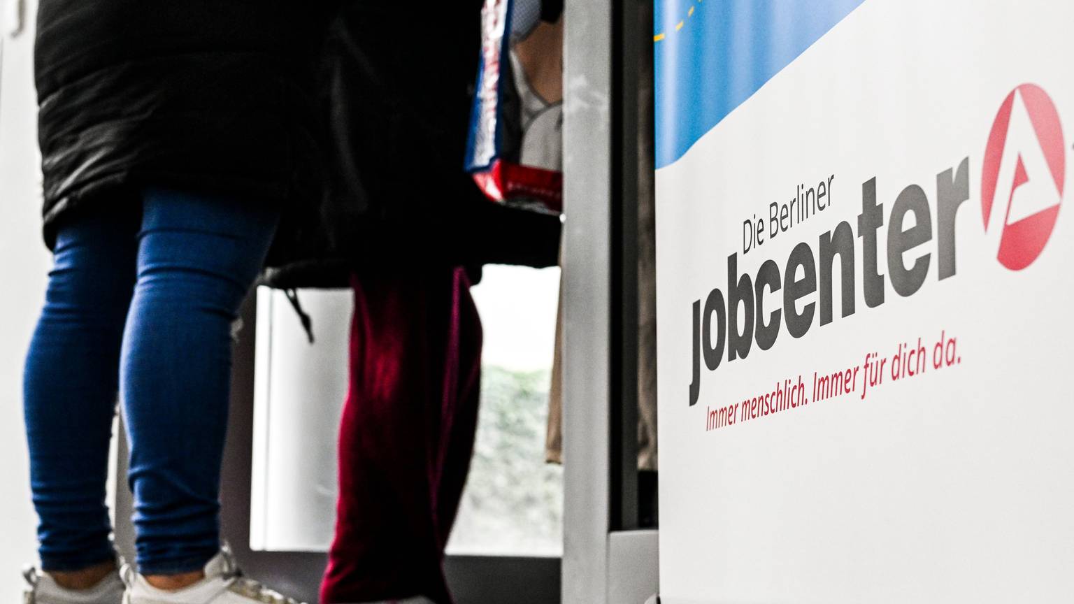 Neben einem Schild, auf dem Jobcenter steht, stehen zwei Personen.
