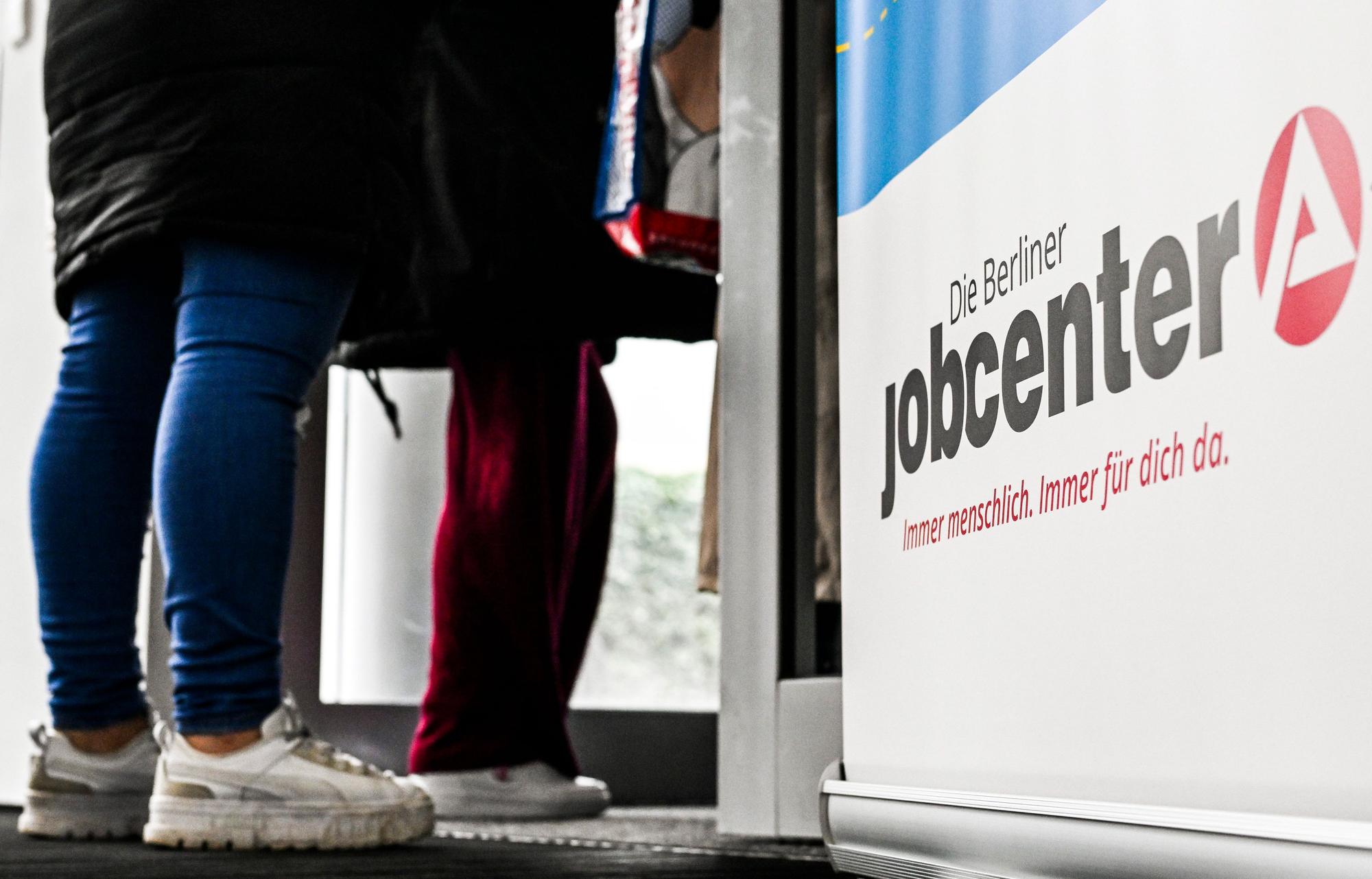 Neben einem Schild, auf dem Jobcenter steht, stehen zwei Personen.