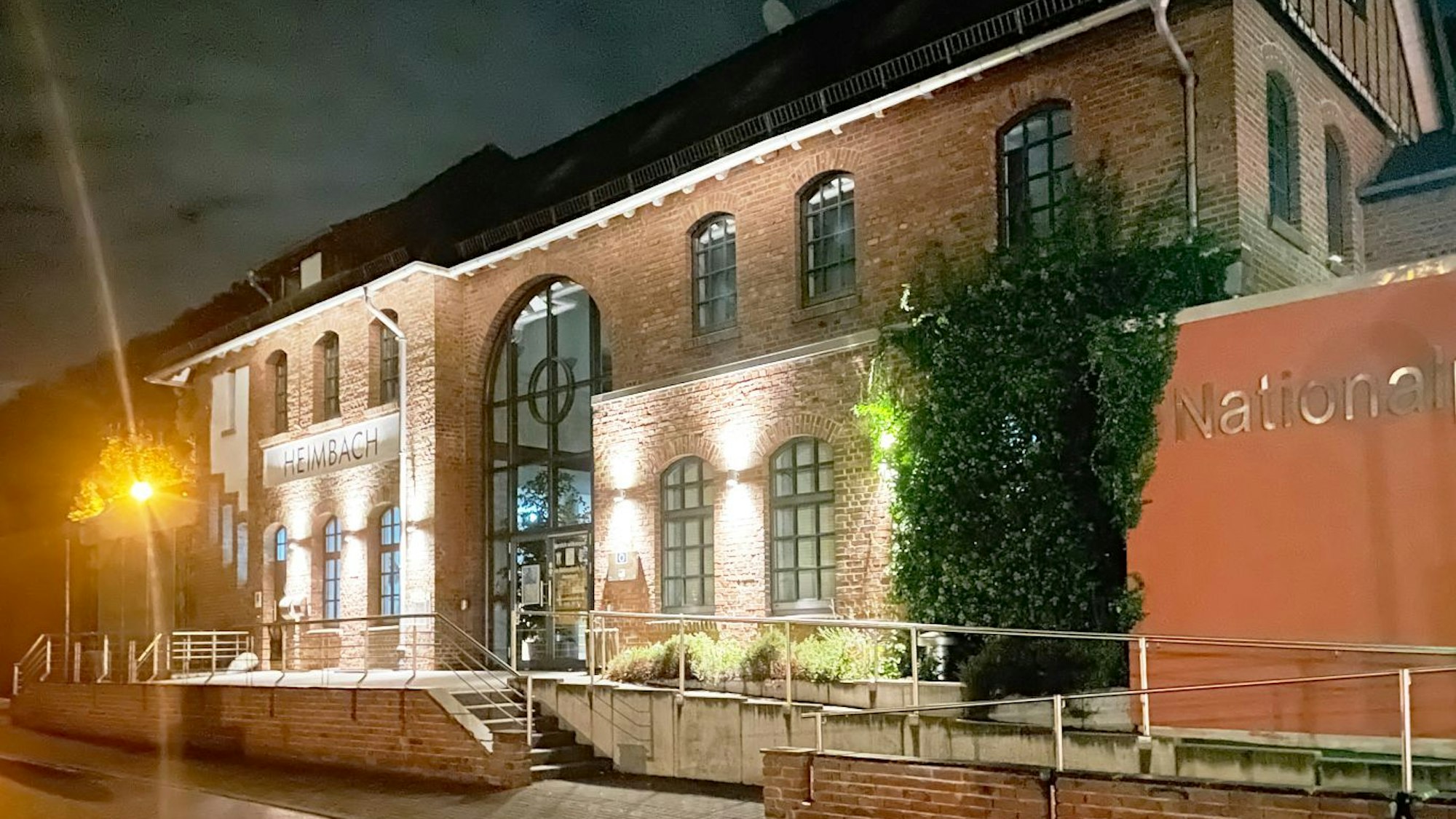 Ein Backsteingebäude wird bei Nacht von kleinen Lampen erleuchtet, unter einer Reihe Fenstern ist ein großes Schild mit der Aufschrift „Heimbach“ zu erkennen.