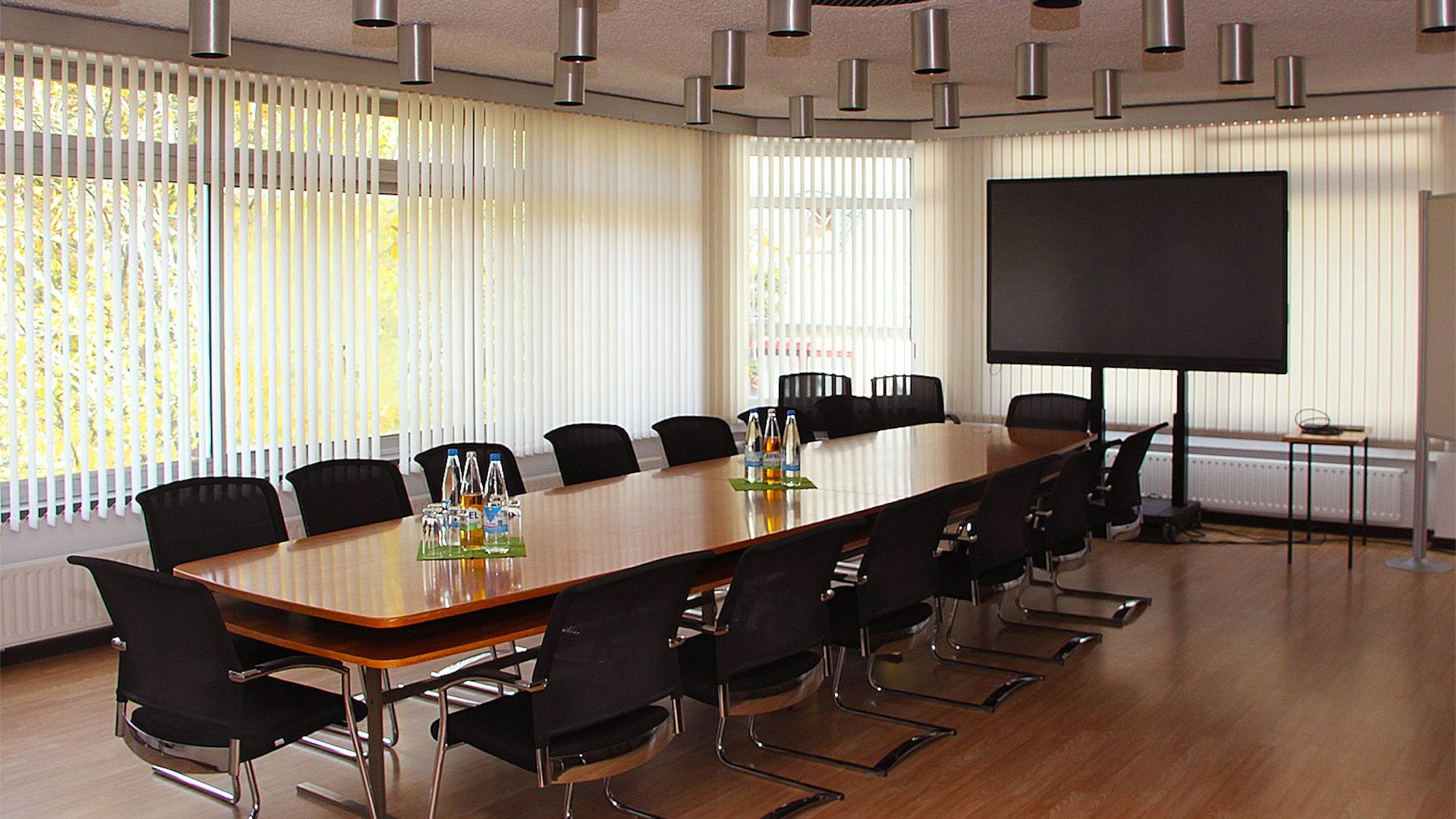In einem Raum mit vielen Fenstern steht ein Tisch aus braunem Holz mit 14 Stühlen, an einer Kopfseite steht ein großer Bildschirm, der ausgeschaltet und daher schwarz ist.