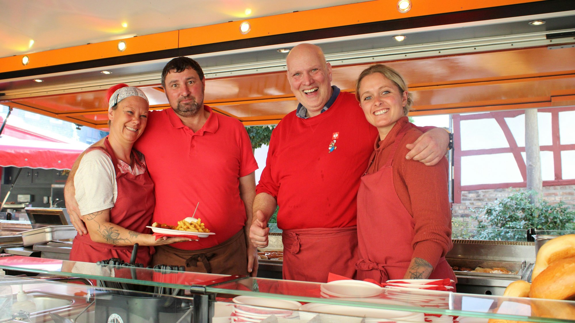 Vier Menschen stehen glücklich in einem Kirmesgeschäft. Eine Frau präsentiert einen Teller mit Pommes und Currywurst.