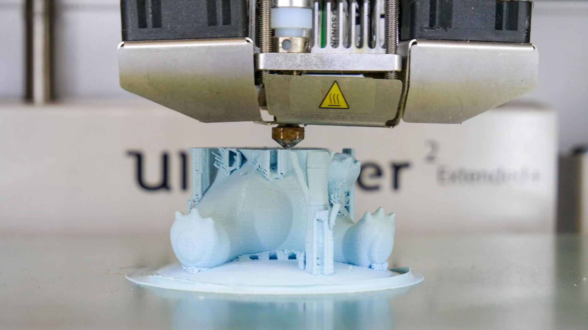 Auf dem Bild ist zu sehen, wie ein 3D-Drucker ein hellblaues Plastikspielzeug herstellt.