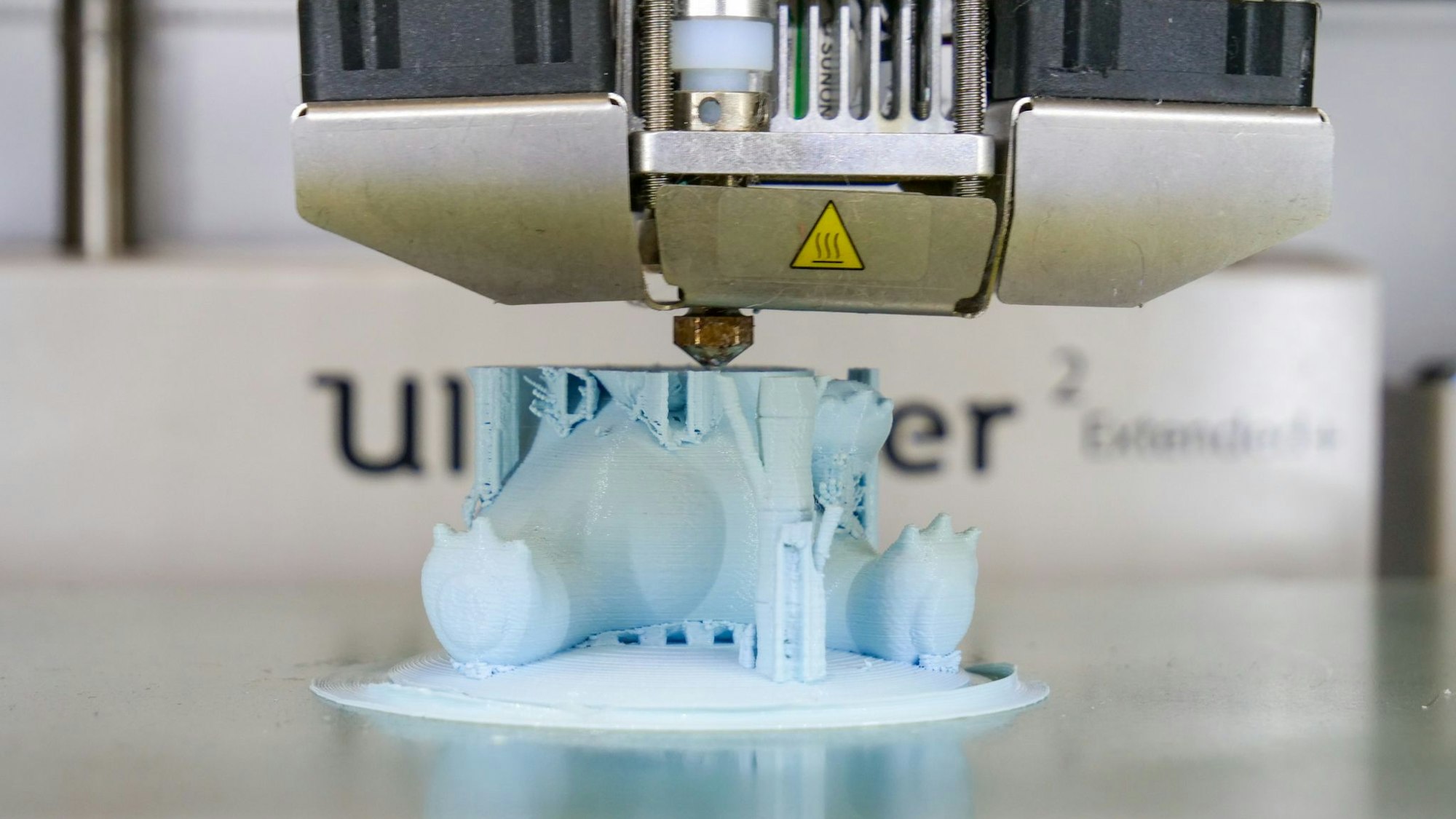 Auf dem Bild ist zu sehen, wie ein 3D-Drucker ein hellblaues Plastikspielzeug herstellt.