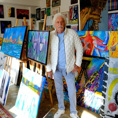 Der Maler Franz Kruse aus Floisdorf steht neben vor einigen bunten Bildern, auf denen Menschen, Pflanzen, aber auch Abstraktes zu sehen sind.