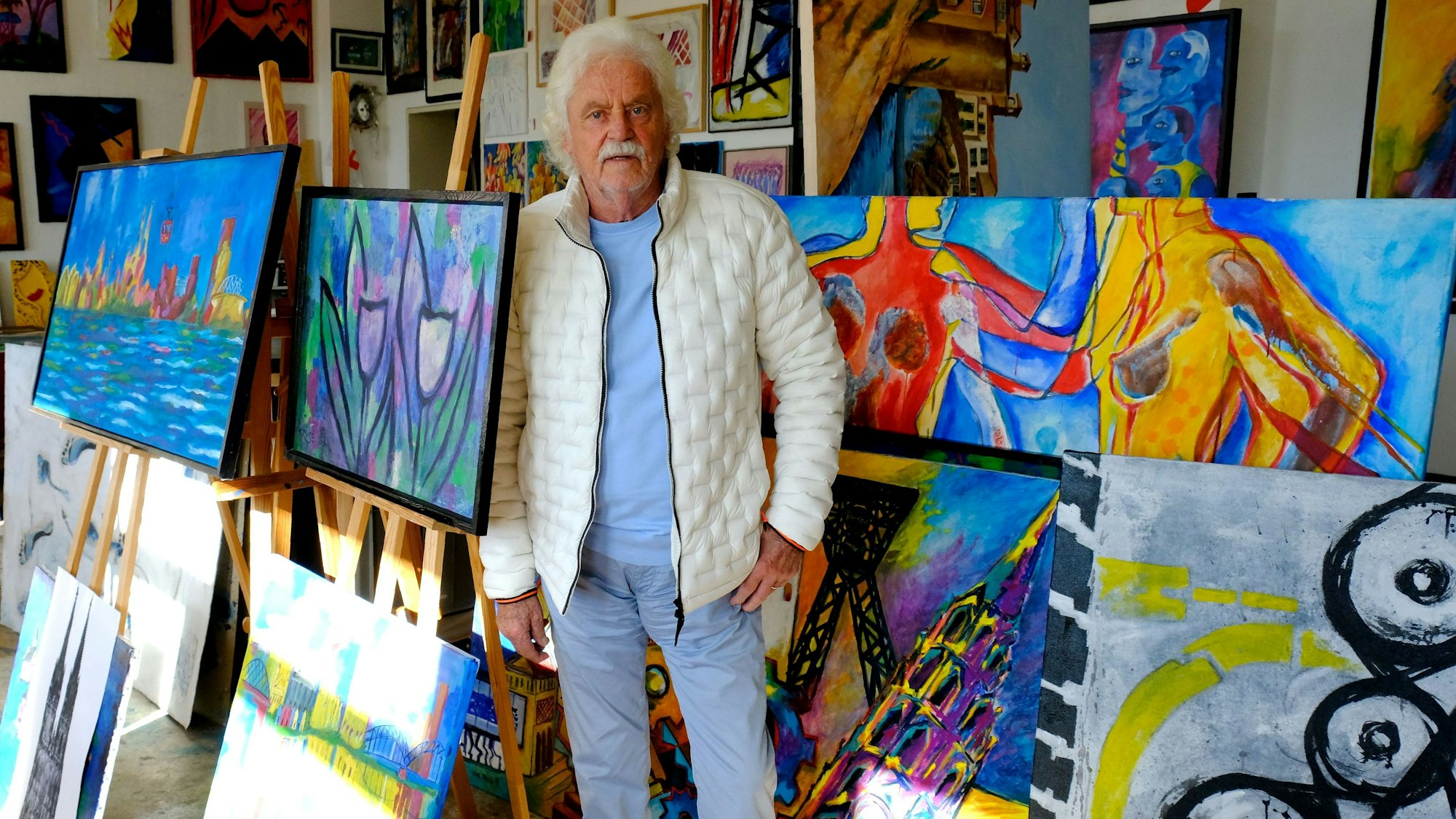 Der Maler Franz Kruse aus Floisdorf steht neben vor einigen bunten Bildern, auf denen Menschen, Pflanzen, aber auch Abstraktes zu sehen sind.