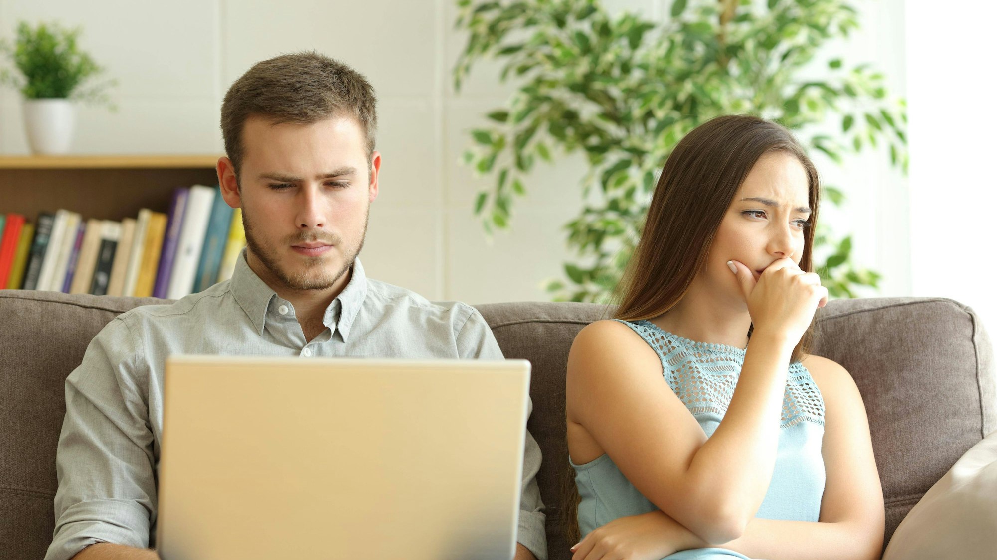 Mann sitzt auf dem Sofa und schaut auf einen Laptop. Daneben sitzt eine Frau, die enttäuscht ins Leere schaut.