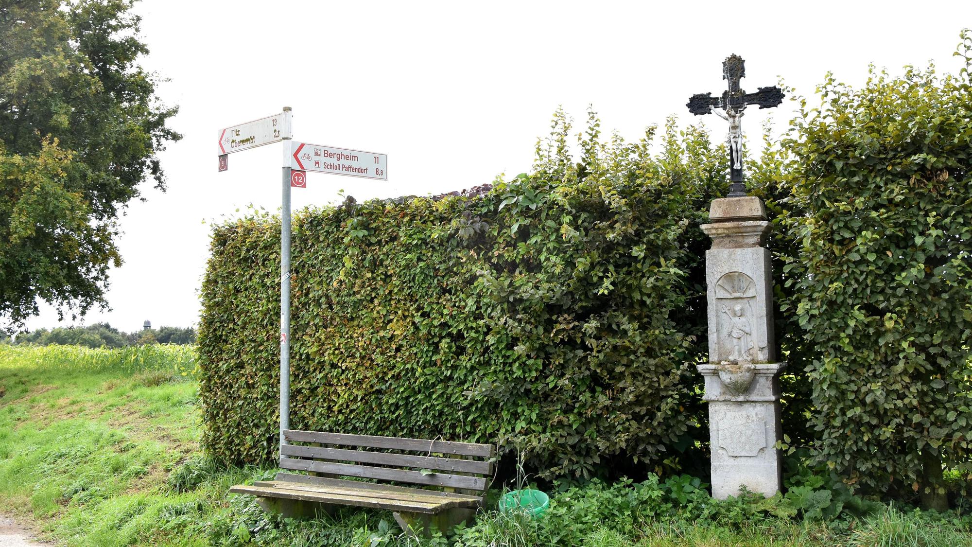 Vor einer Hecke stehen eine Bank und ein Kreuz.