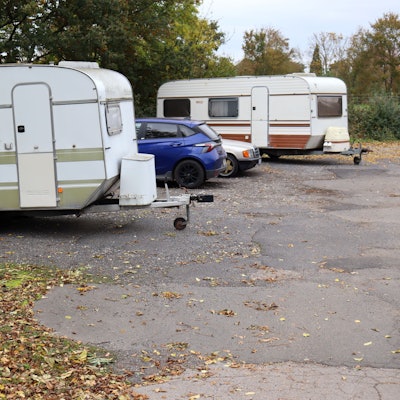 Auf einem Park-and-Ride-Platz stehen zwei Wohnwagenanhänger und zwei Autos. Auf dem Boden liegt Laub.
