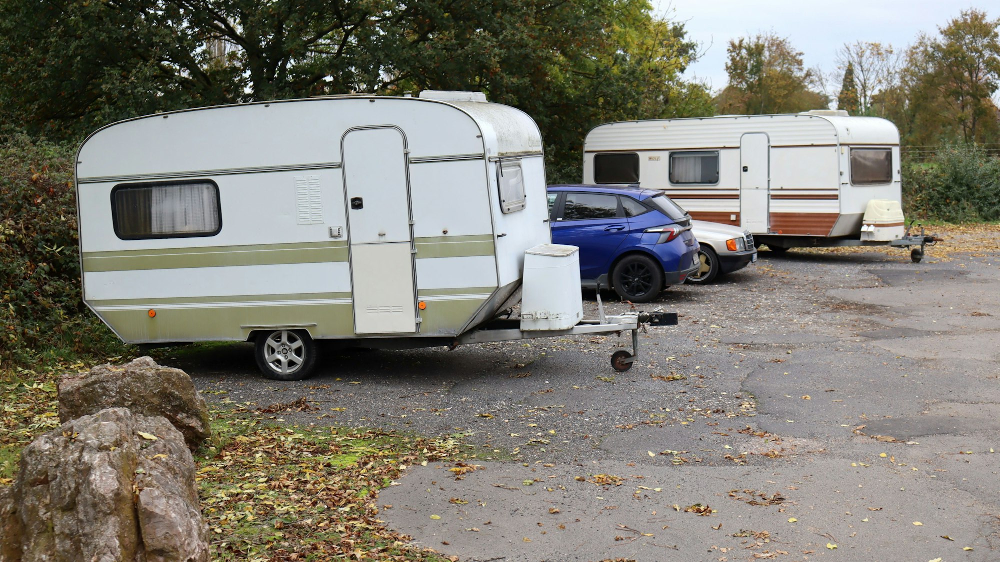 Auf einem Park-and-Ride-Platz stehen zwei Wohnwagenanhänger und zwei Autos. Auf dem Boden liegt Laub.