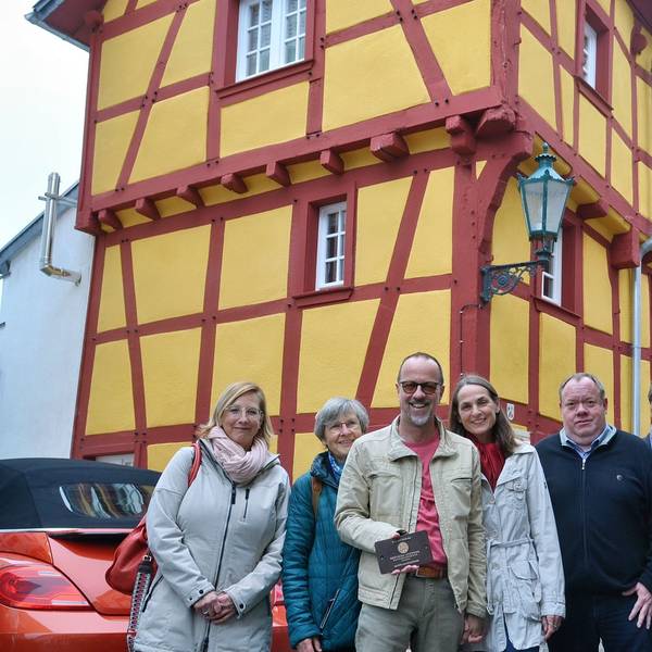 Eine Gruppe Menschen steht vor einem Fachwerkhaus mit gelben Gefachen und roten Balken.
