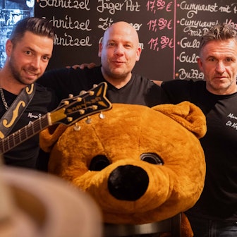 Das Bild zeigt die drei Organisatoren, die sich in den Arm nehmen. Vor Jörg Peter liegt ein überdimensionaler Plüschbärenkopf.