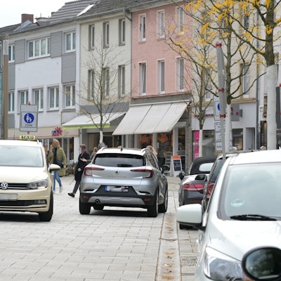 Am Straßenrand parken Autos. In der Mitte der Fußgängerzone begegnen sich zwei Autos.