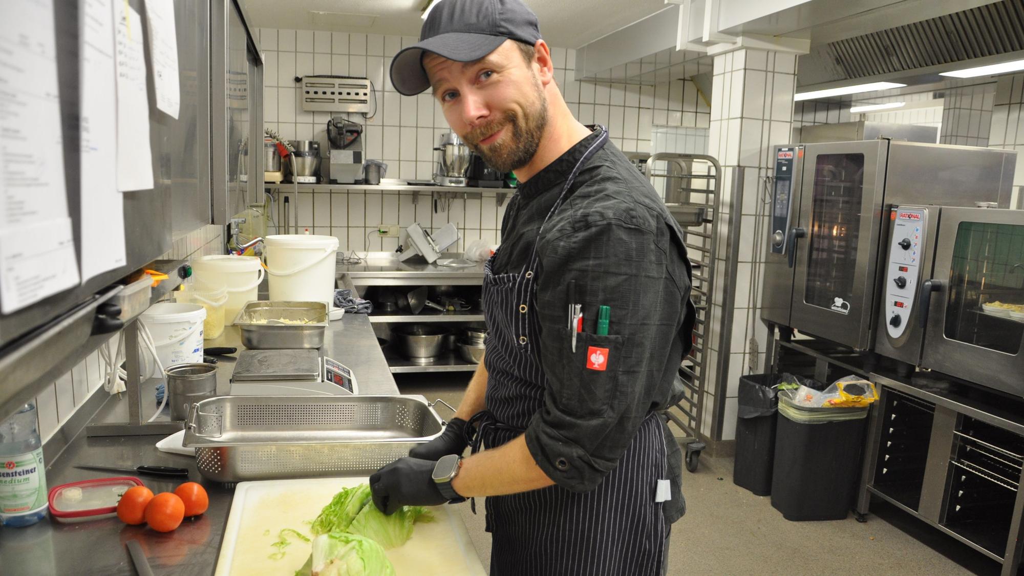 Der Koch trägt schwarze Kleidung, eine Mütze und Handschuhe, während er in der Kantine Salat schnippelt.