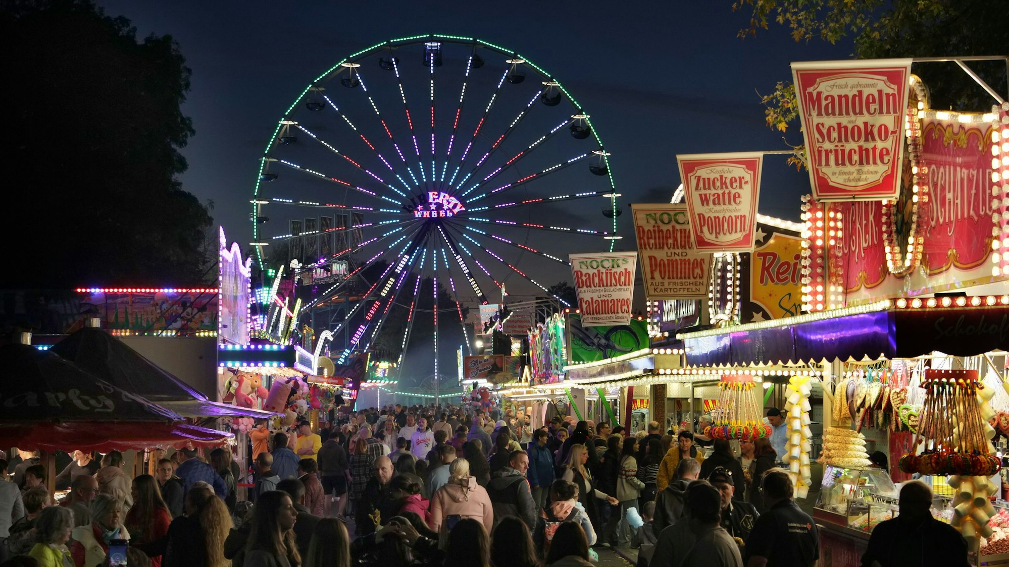 Eine Menschenmenge besucht die beleuchteten Stände, in denen Zuckerwatte, Backfisch und Popcorn verkauft werden.
