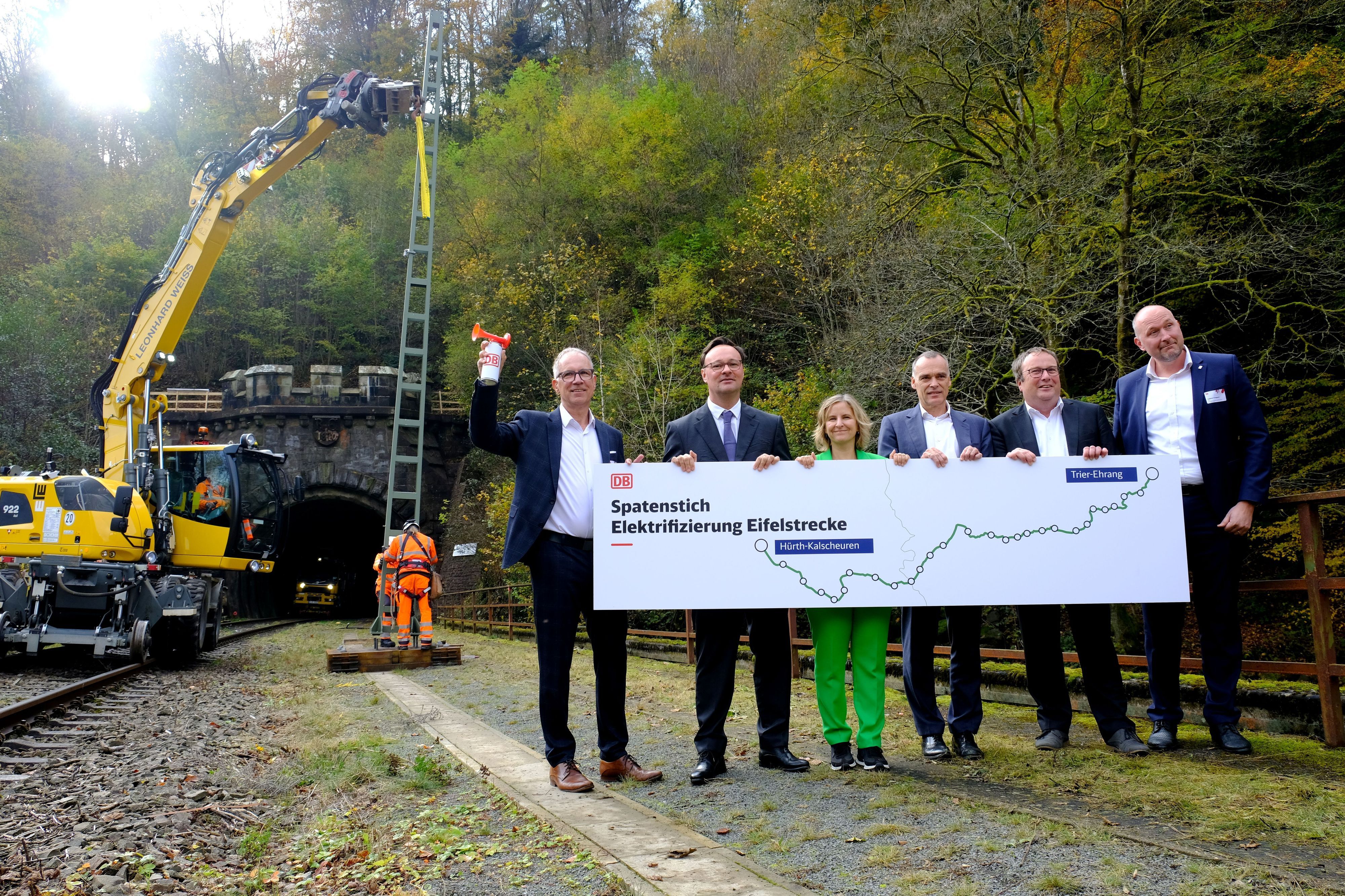 Politiker halten gemeinsam ein Transparent mit der Aufschrift „Spatenstich Elektrifizierung Eifelstrecke“. Im Hintergrund richten Arbeiter einen Oberleitungsmast auf.