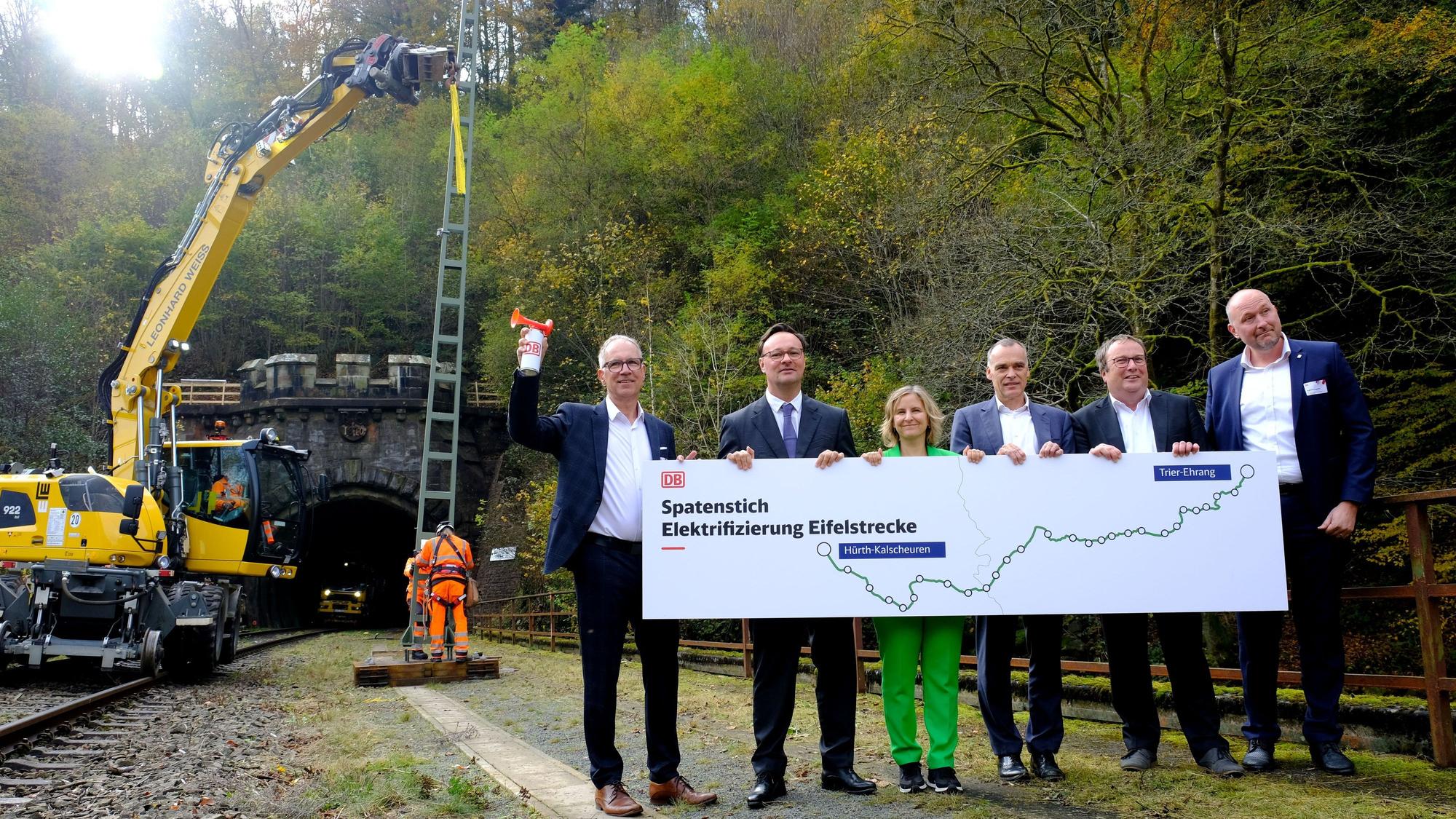Politiker halten gemeinsam ein Transparent mit der Aufschrift „Spatenstich Elektrifizierung Eifelstrecke“. Im Hintergrund richten Arbeiter einen Oberleitungsmast auf.