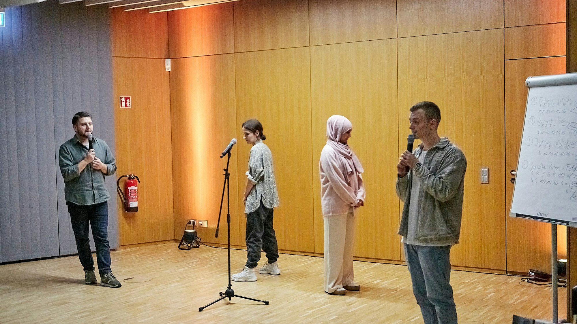Zwei Frauen, die einander den Rücken zuwenden, und zwei Männer mit Mikrofonen stehen in einem Raum.
