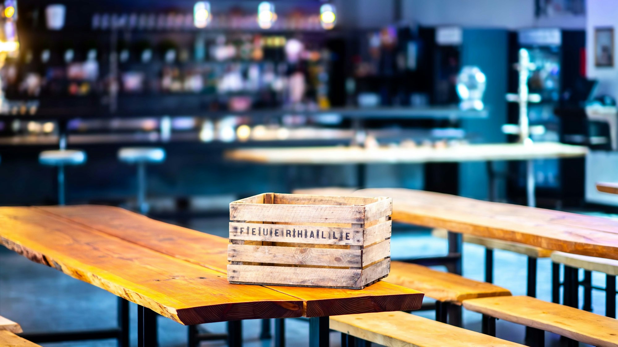 Das Bild zeigt eine leere Holzkiste auf einem Tisch. Im Hintergrund ist ein Teil der Feuerhalle zu sehen.