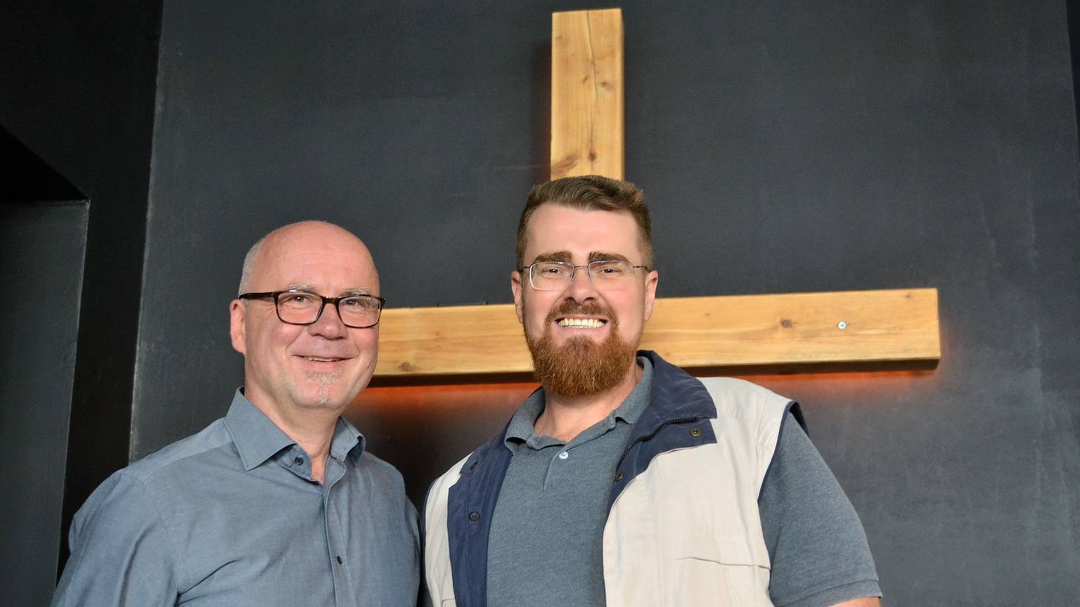 Eljoenai Wildemann und Markus Pfitzer stehen in einem Raum der Freien Christengemeinde Euskirchen vor einem Holzkreuz.