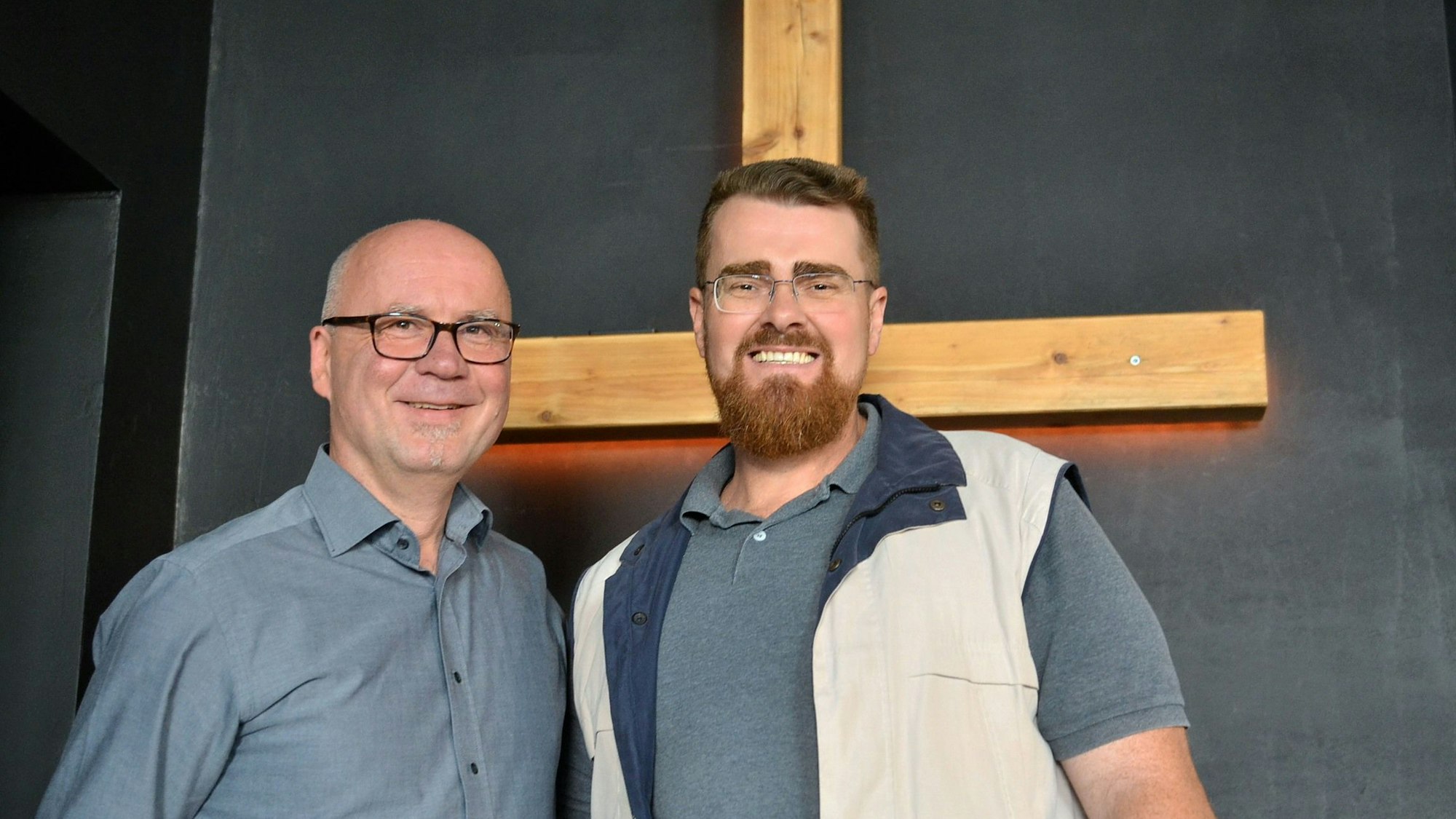 Eljoenai Wildemann und Markus Pfitzer stehen in einem Raum der Freien Christengemeinde Euskirchen vor einem Holzkreuz.