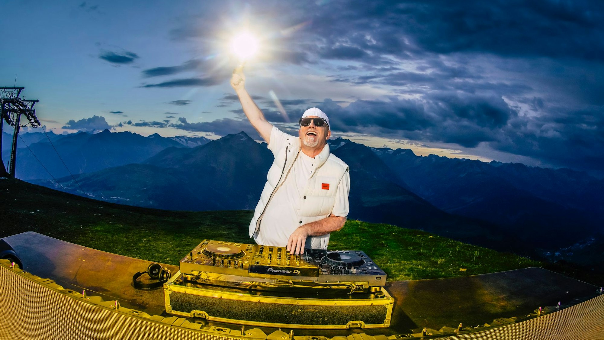 DJ Ötzi steht vor einem DJ-Pult. Hinter ihm sind Berge zu sehen.