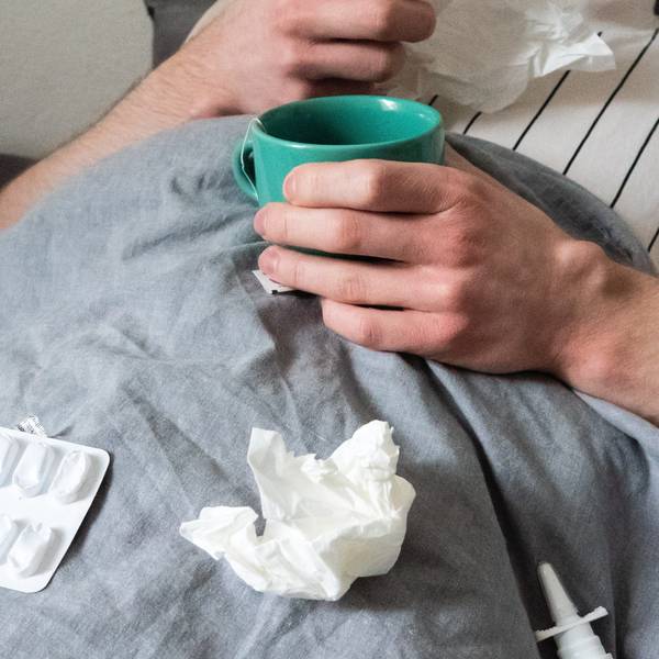 Ein Mann liegt mit Taschentüchern, Teetasse, Nasenspray und Tabletten im Bett.