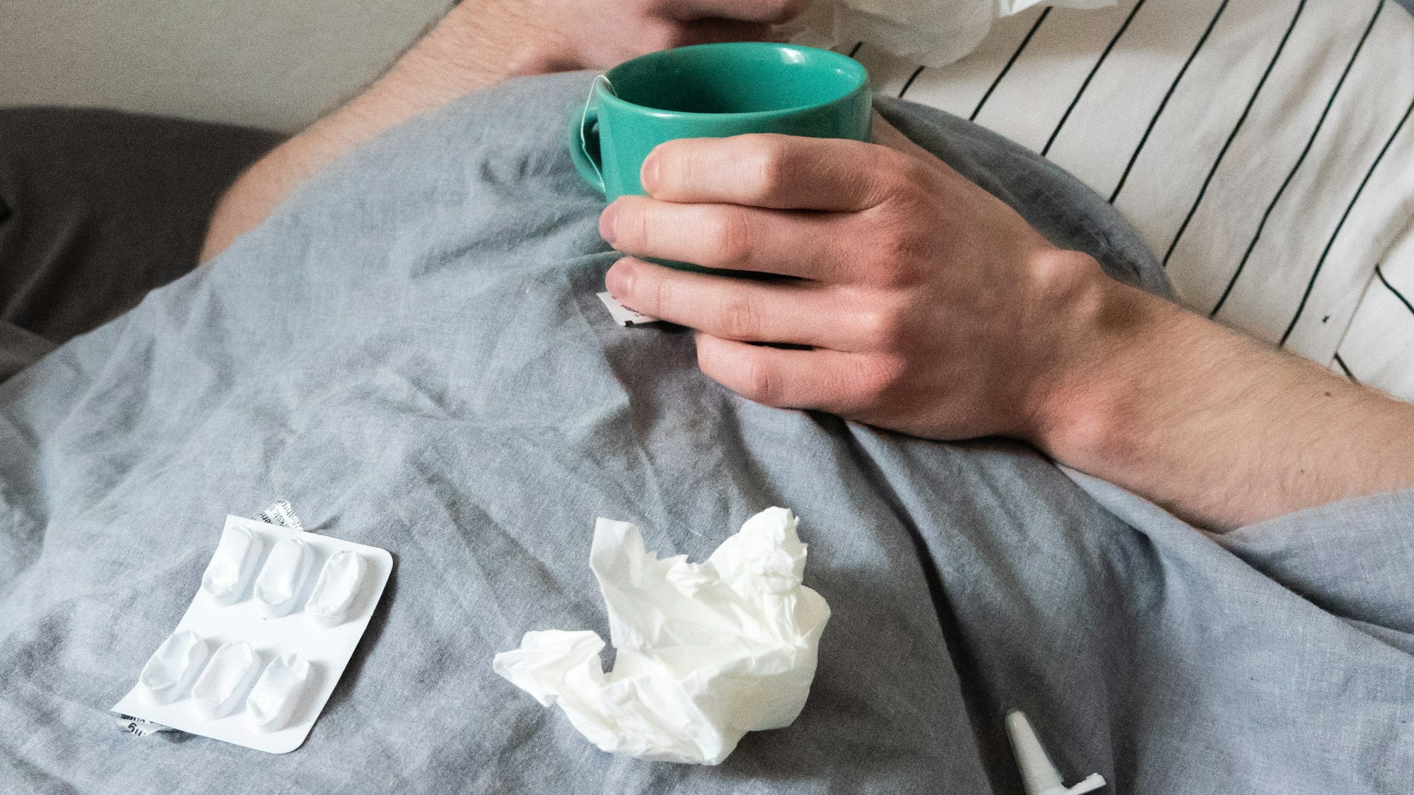 Ein Mann liegt mit Taschentüchern, Teetasse, Nasenspray und Tabletten im Bett.