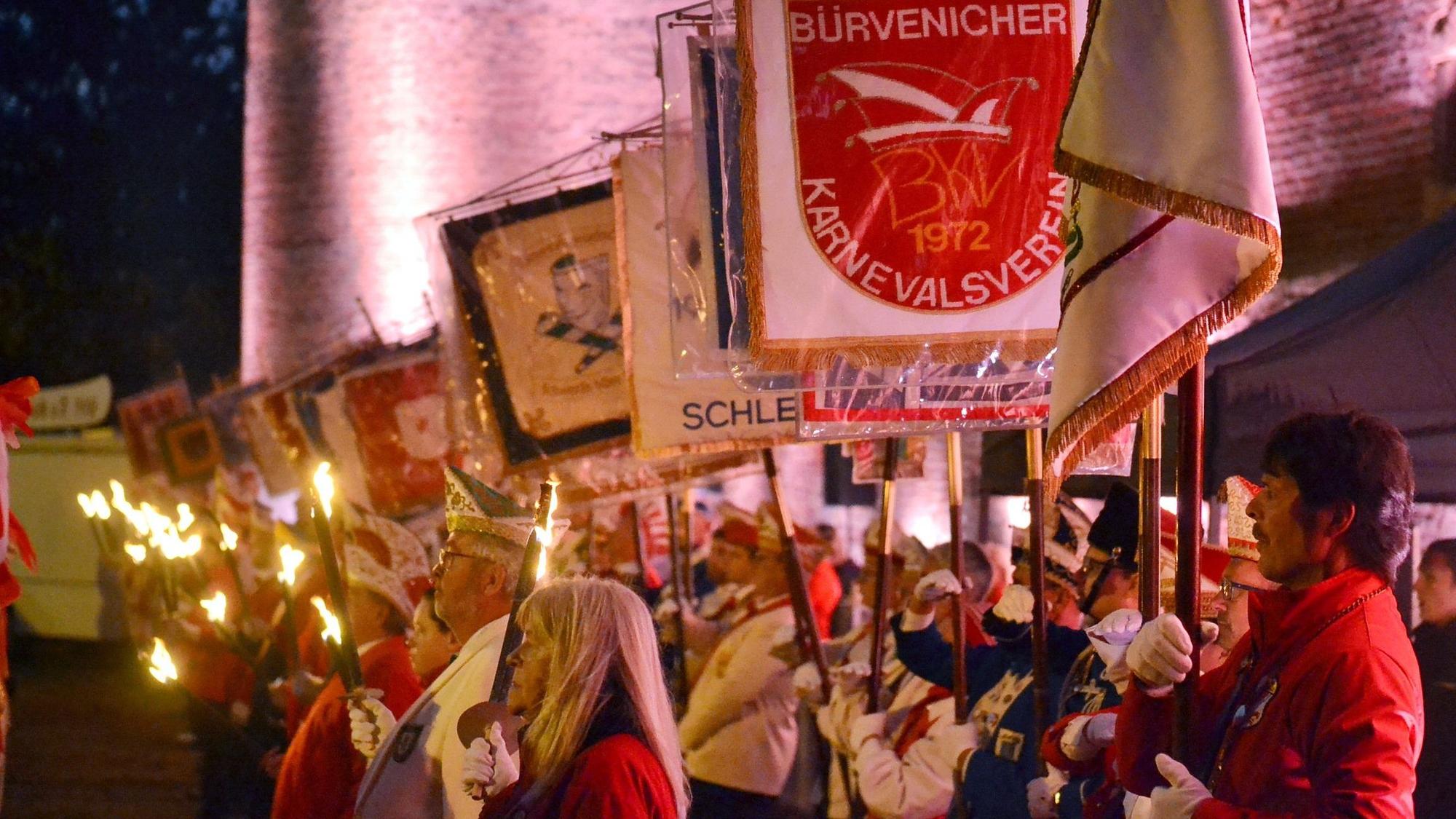 Zahlreiche Karnevalisten in roten und weißen Jacken stehen beim Zapfenstreich in Zülpich an einer Mauer. Sie halten die Fahnen ihrer Vereine oder Pechfackeln in den Händen.