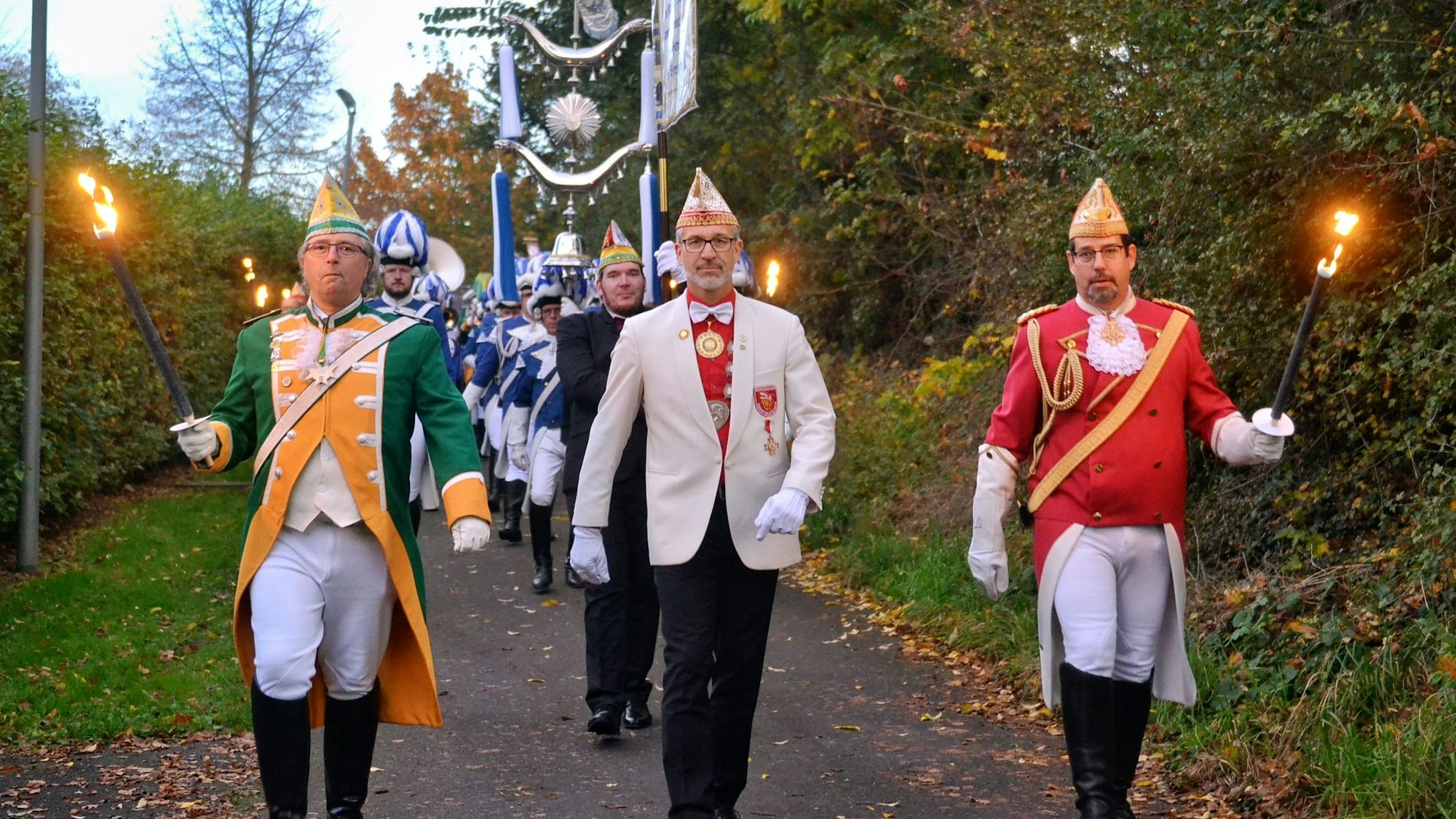 Drei Männer in den Uniformen ihrer Karnevalsvereine gehen an der Spitze eines Umzugs in Zülpich, zwei von ihnen halten Pechfackeln in den Händen. Hinter ihnen sind zahlreiche Musiker zu erkennen.