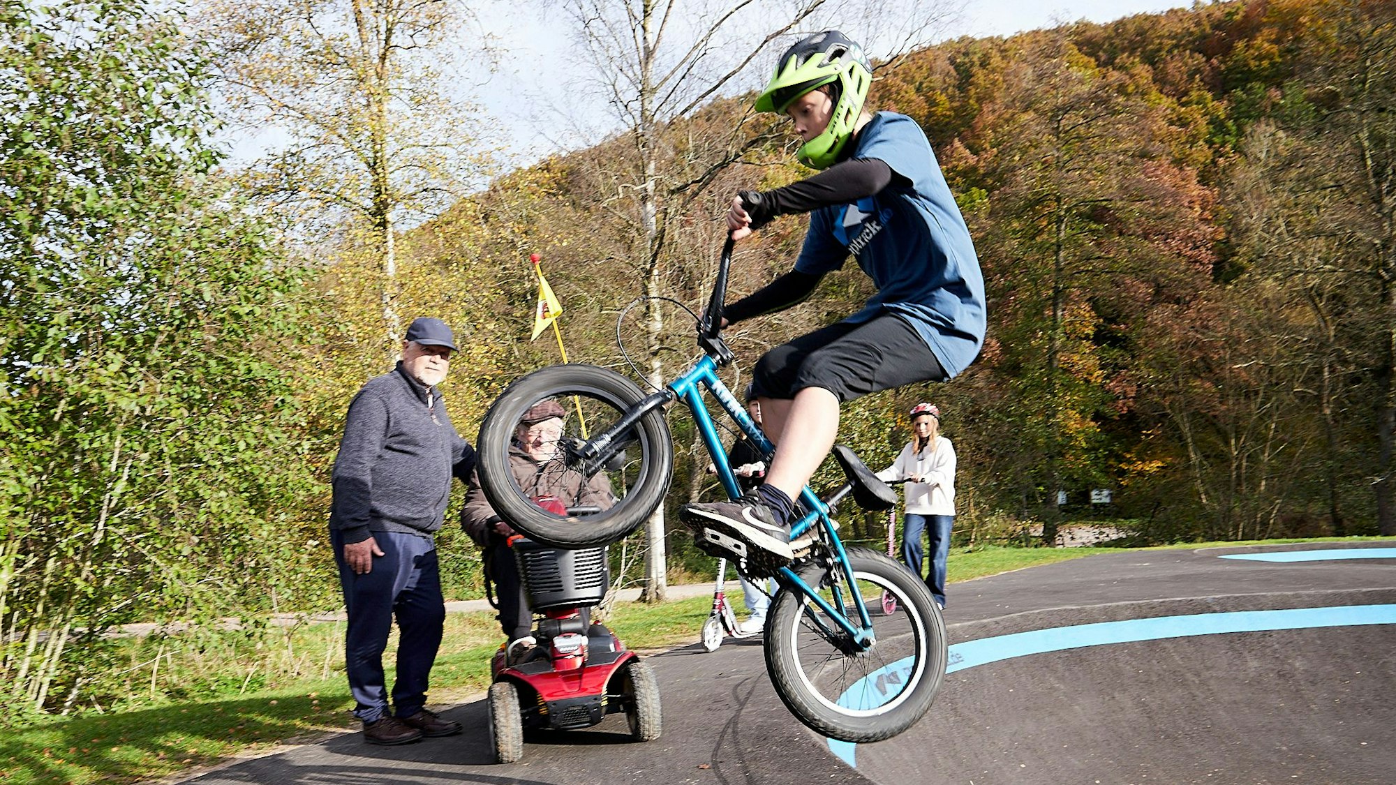 Ein BMX-Fahrer setzt an einem Pumptrack zum Sprung an und wird dabei von zwei älteren Herren beobachtet. Einer sitzt in einem E-Chopper.