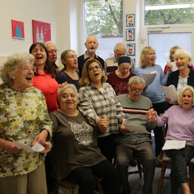 Frauen und Männer singen, einige sitzen, andere stehen. Einige halten sich an den Händen.