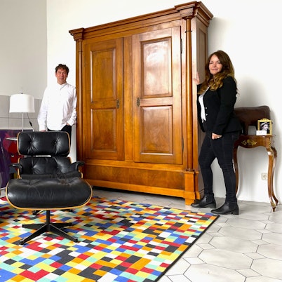 Thomas Dyck mag moderne Designerklassiker, Linda Brovot liebt antike Möbel. Beide lehnen an einem Empire Schrank von ca. 1800, im Vordregrund steht ein Eames Lounge Chair aus dem Jahre 1956, auf dem Fußboden liegt ein „1024 Farben Gerhard Richter Teppich“.