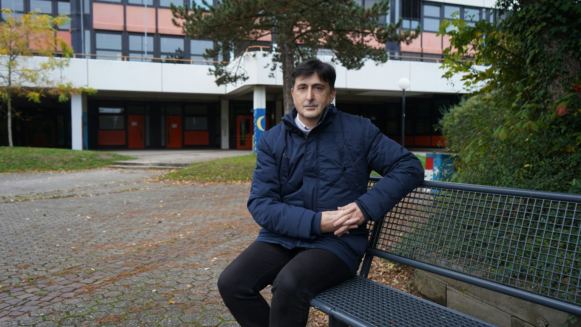 Das Foto zeigt einen Mann, der auf einer Bank vor einem Schulblock sitzt.