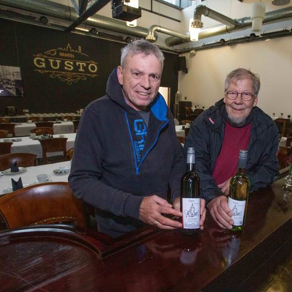 Zwei Männer stehen am Tresen einer Gaststätte und halten zwei Flaschen mit Wein in Händen.