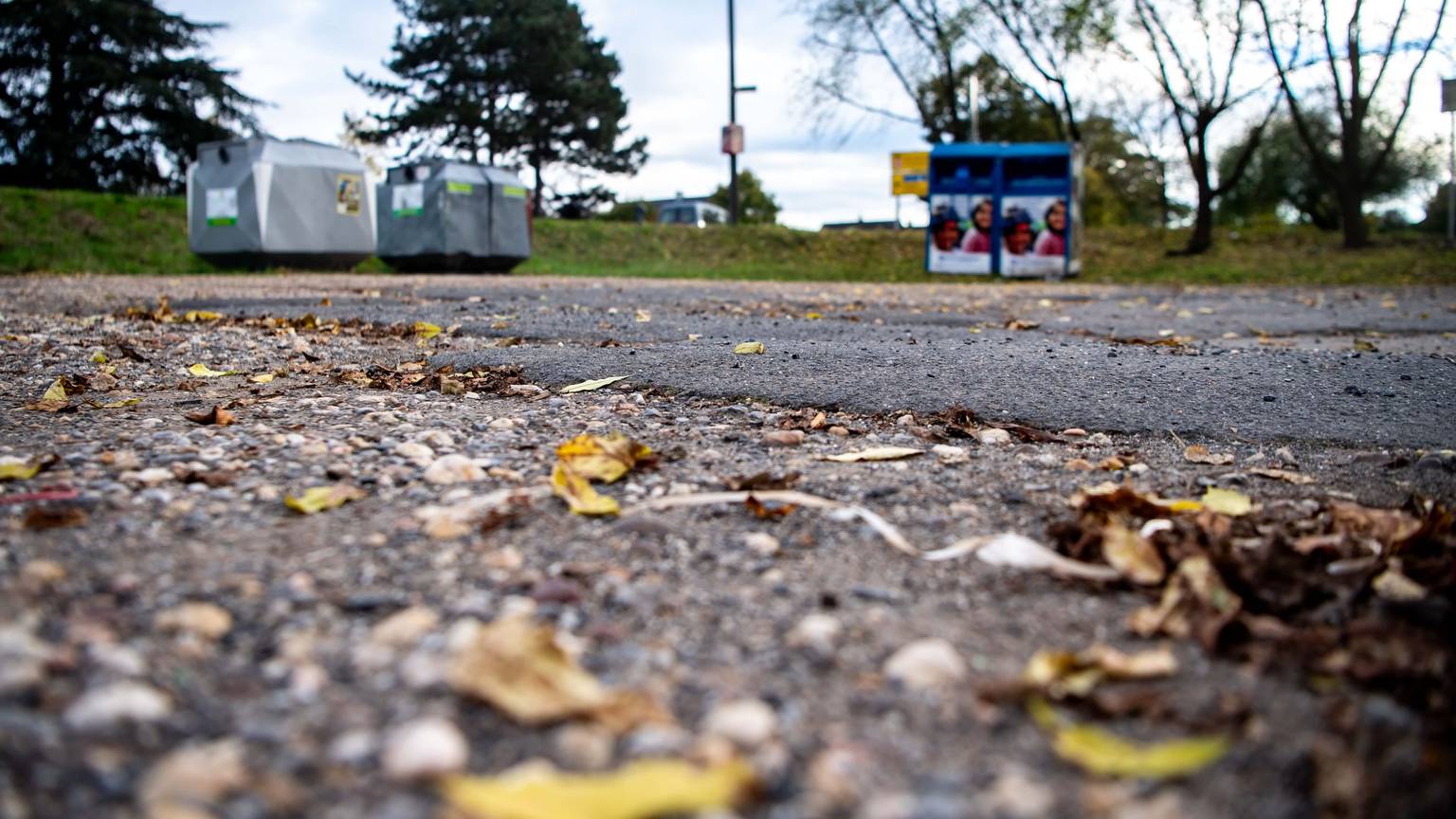 Blätter liegen auf einem Parkplatz. Die Asphaltdecke ist geflickt. Im Hintergrund stehen Leergut- und Altkleidercontainer.